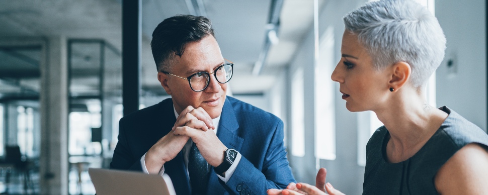 Ein Mann und eine Frau im Geschäftsoutfit unterhalten sich in einem Büro. 