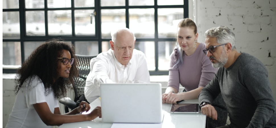 Eine Gruppe von vier Fachkräften unterschiedlichen Alters sitzt zusammen und schaut auf einen Laptop-Bildschirm, was ein altersinkludierendes Arbeitsumfeld veranschaulicht.