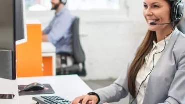 Person im Büro mit Headset am Computer, lächelnd.
