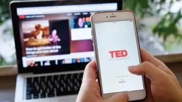 Person hält Smartphone mit TED-App, im Hintergrund ein Laptop.