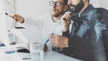 Zwei Personen arbeiten am Computer, eine zeigt auf den Bildschirm.