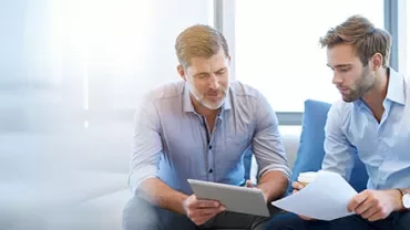 Zwei Personen diskutieren in einem Büro mit einem Tablet und Dokumenten.