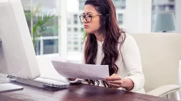 Person im Büro liest papierbasierte Unterlagen am Computer.