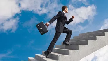 Person im Anzug geht eine Treppe hinauf unter blauem Himmel.