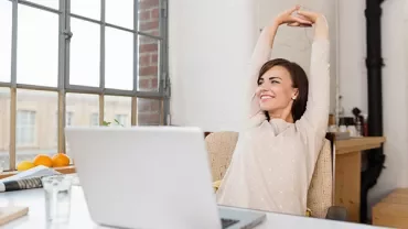 Person in heller Kleidung streckt sich am Schreibtisch mit Laptop.