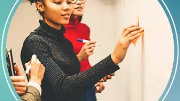 Person schreibt mit Filzstift auf ein Whiteboard, andere schauen zu.