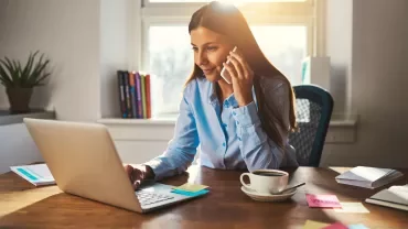 Person telefoniert und arbeitet am Laptop an einem Schreibtisch mit Kaffeetasse.