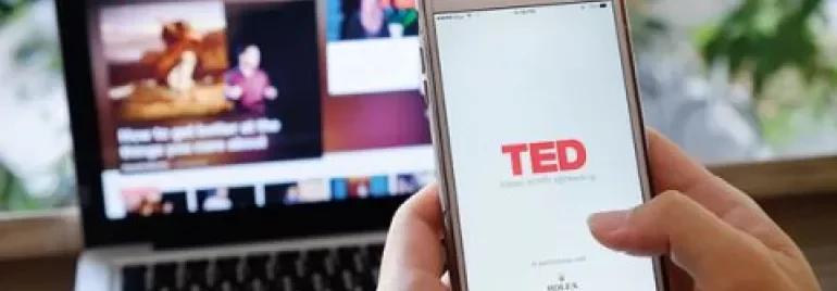 Person hält Smartphone mit TED-App, im Hintergrund ein Laptop.