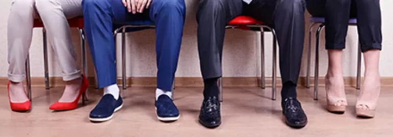 Vier Menschen sitzen nebeneinander auf Stühlen, Fokus auf Beine und Schuhe.