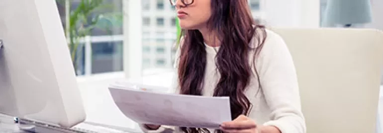 Person im Büro liest papierbasierte Unterlagen am Computer.
