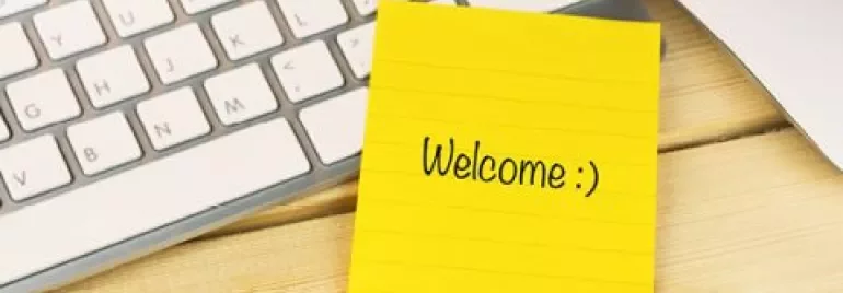 Gelbe Notiz mit 'Welcome :)', Tastatur und Stift auf Schreibtisch.