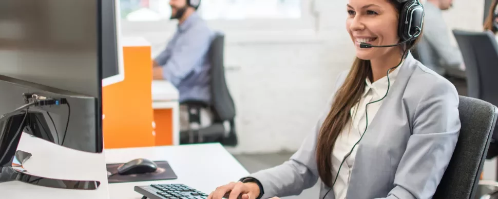 Person im Büro mit Headset am Computer, lächelnd.