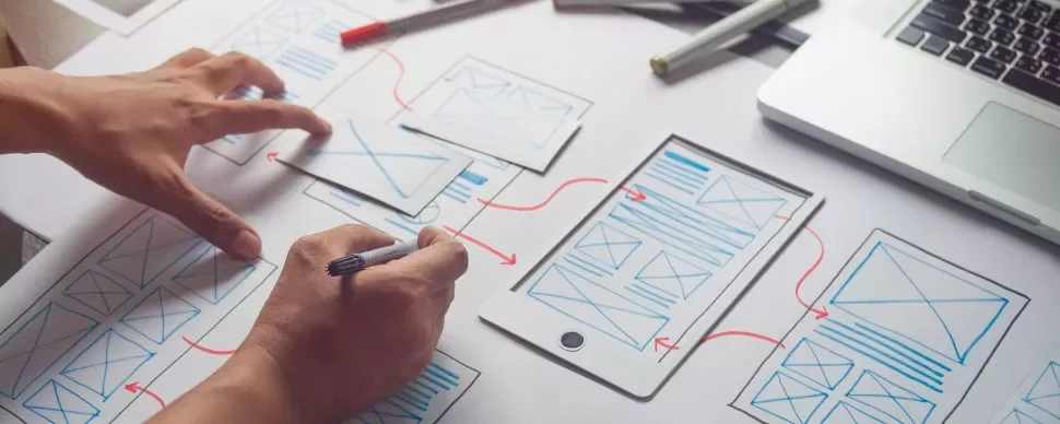 Person zeichnet Wireframe-Designs auf Papier mit einem Tablet daneben.