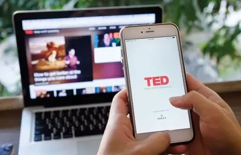 Person hält Smartphone mit TED-App, im Hintergrund ein Laptop.