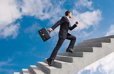Person im Anzug geht eine Treppe hinauf unter blauem Himmel.