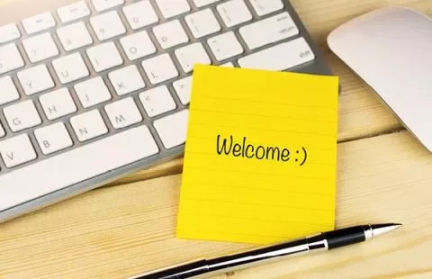 Gelbe Notiz mit 'Welcome :)', Tastatur und Stift auf Schreibtisch.