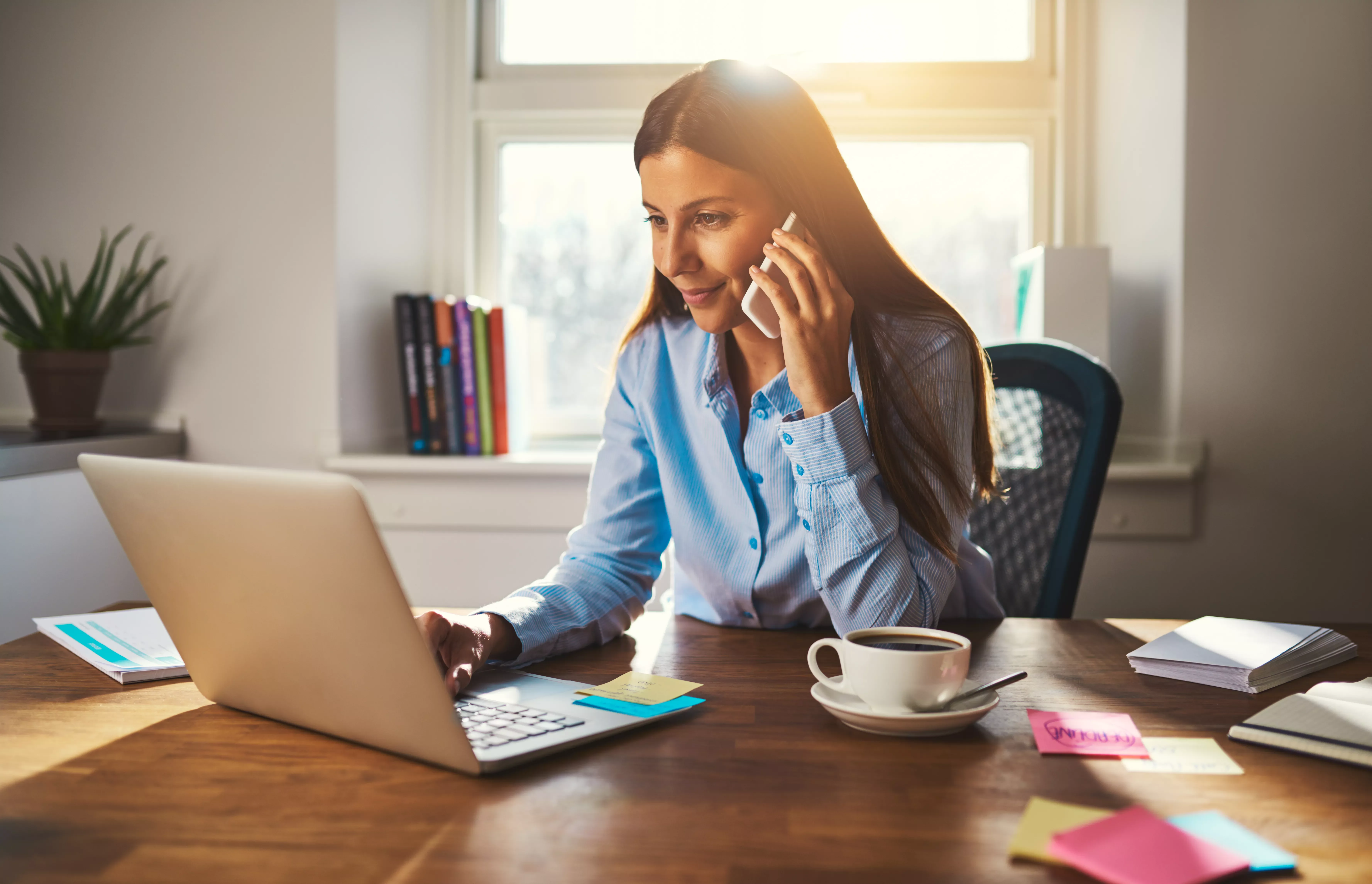 Person telefoniert und arbeitet am Laptop an einem Schreibtisch mit Kaffeetasse.
